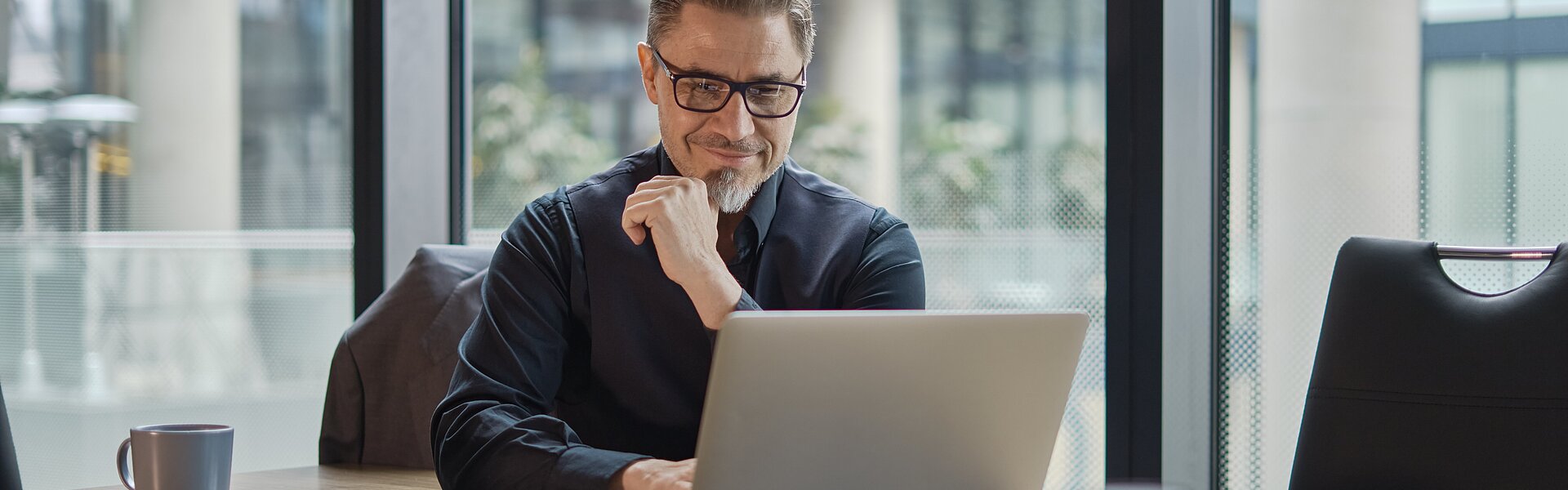 Finanzbuchhalter arbeitet am Laptop im Büro mit großer Fensterfront im Hintergrund
